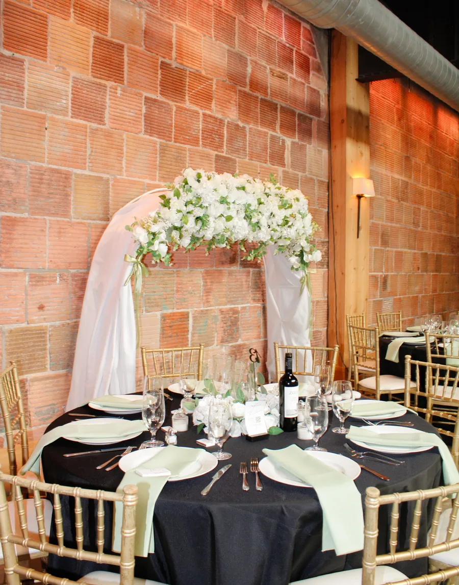 A white floral arch draped in fabric over a round table with a black cloth, against a brick wall