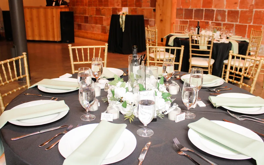 A round table with a black cloth, sage napkins, white flowers and candles, brick wall and gold chairs behind