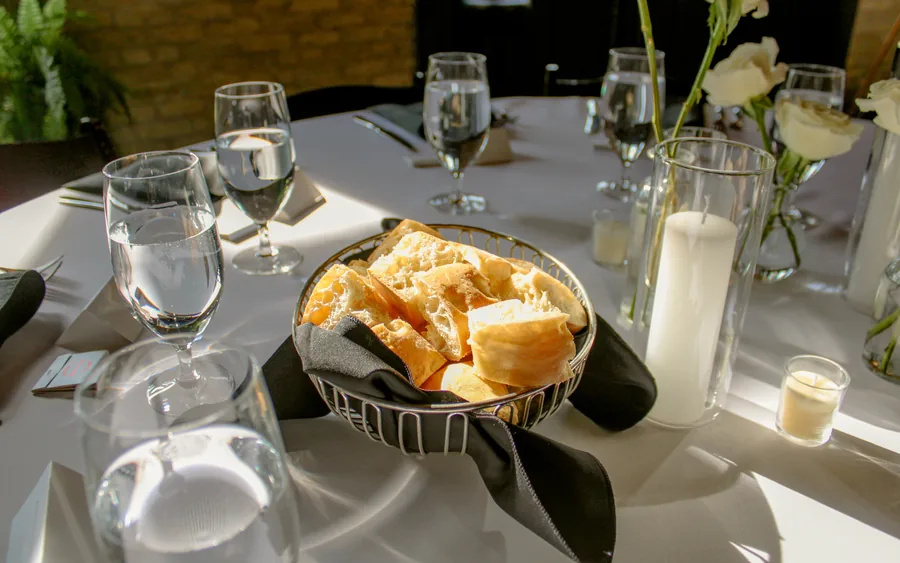 A basket of bread on a white tablecloth beside water glasses and a pillar candle