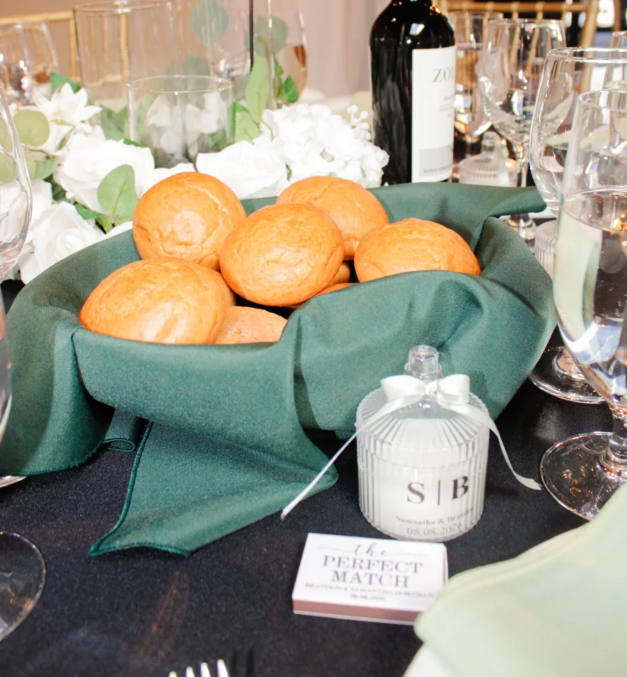 A basket of bread rolls in a green napkin beside white flowers and a favor box on a black tablecloth