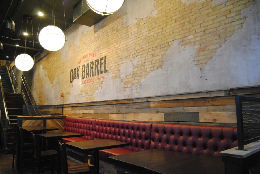 The painted Oak Barrel sign on a cream brick wall above a red tufted banquette and wooden tables