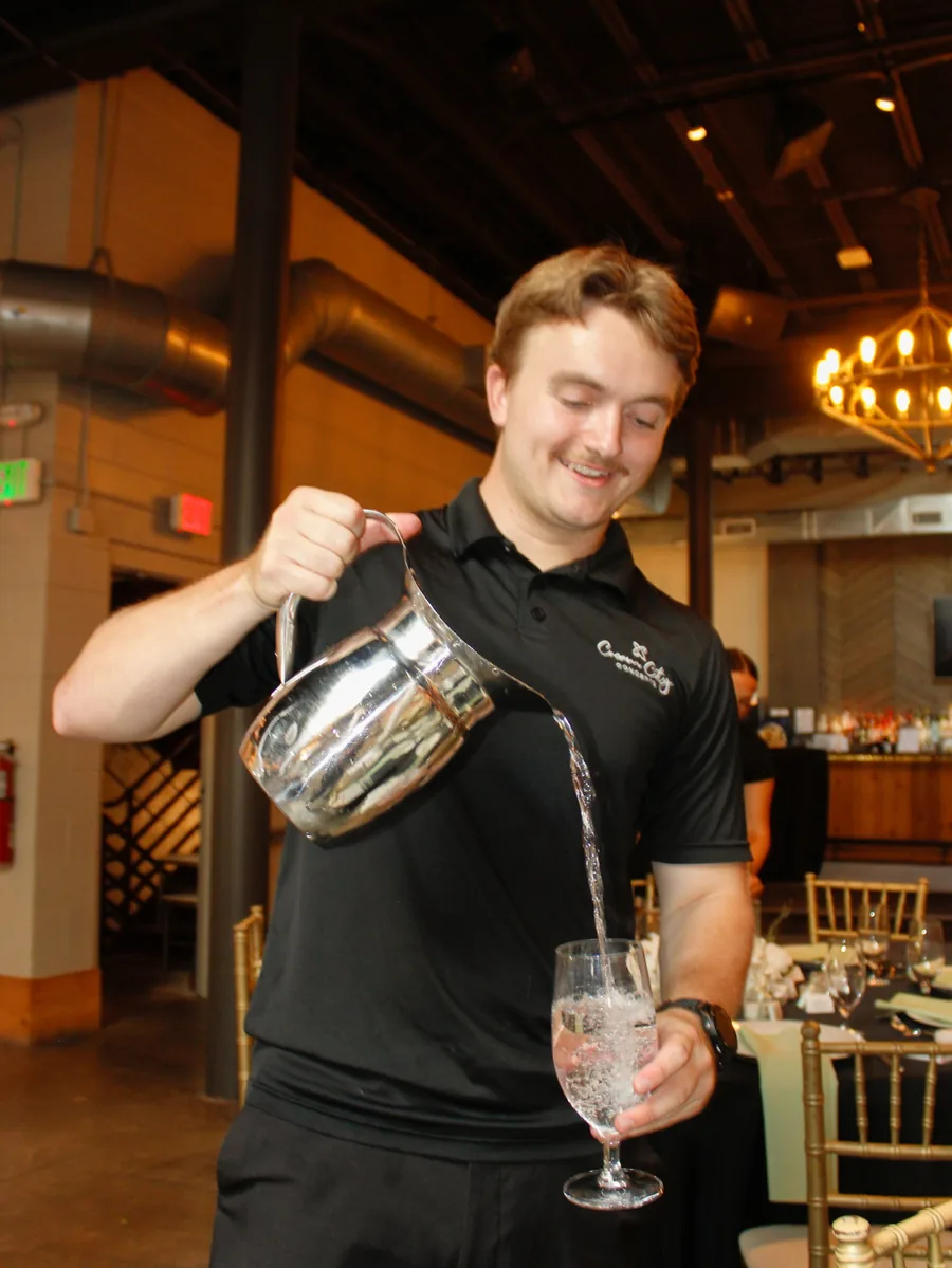 A server in a black polo pouring water from a silver pitcher into a stemmed glass in a room with a brass chandelier