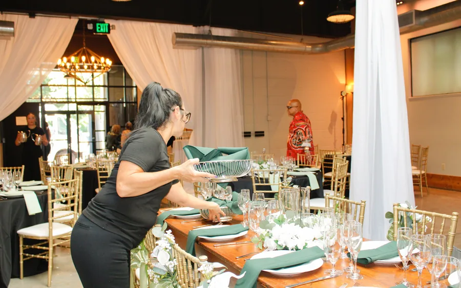 A server in black setting a green napkin basket on a long wooden table under white drapes and a chandelier